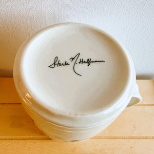 Op de afbeelding: Een witte keramische mok met een oor, van onderaf gezien. De bodem van de mok heeft een handtekening in zwart schrift: "Starla M. Halfmann". De mok staat op een lichtbruin houten oppervlak.