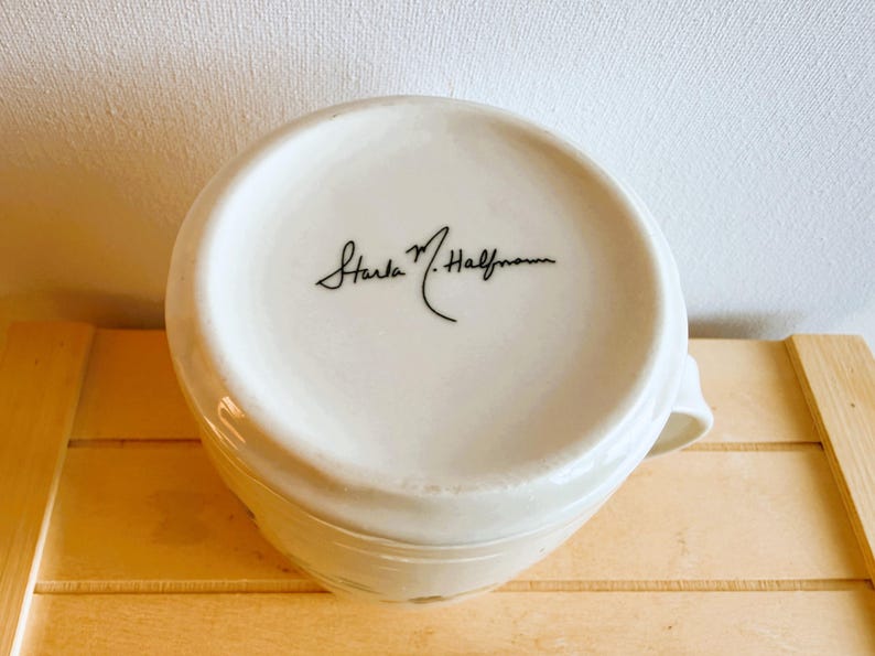 Op de afbeelding: Een witte keramische mok met een oor, van onderaf gezien. De bodem van de mok heeft een handtekening in zwart schrift: "Starla M. Halfmann". De mok staat op een lichtbruin houten oppervlak.