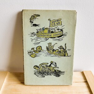 May include: Vintage hardcover book titled "Nonsense Songs" by Edward Lear. The cover features whimsical illustrations in black and yellow, including a boat with characters, a fish, and a turtle. The book is set on a wooden surface.
