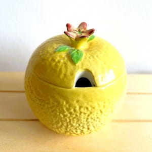 Puede incluir: Un azucarero de cerámica amarillo con forma de limón con una hoja verde y una flor roja en la parte superior. El tazón tiene un pequeño pico para verter.