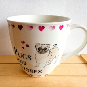 May include: White ceramic mug with a pug illustration and the words "Pugs and Kisses." The mug features pink and maroon heart accents around the rim and base. The mug is sitting on a wooden surface.