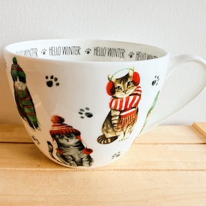 Könnte beinhalten: Ein weißer Keramikbecher mit Illustrationen von Katzen, die Wintermützen, Schals und Ohrenschützer tragen, akzentuiert durch schwarze Pfotenabdrücke. Der Innenrand ist mit dem wiederholten Text HELLO WINTER in schwarzer Schrift bedruckt.