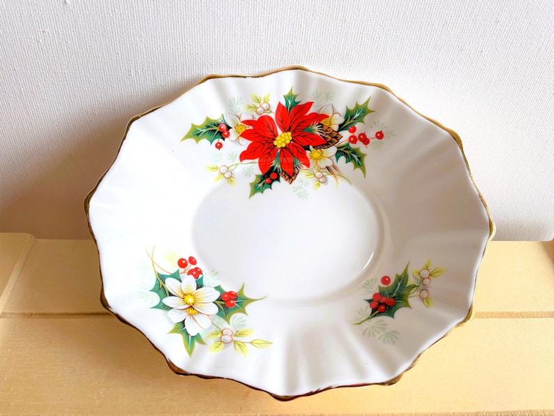 K&ouml;nnte beinhalten: Eine wei&szlig;e Keramikschale mit goldfarbenem Rand zeigt ein weihnachtliches Design. Die Schale ist mit roten Weihnachtssternen, gr&uuml;nen Stechpalmenbl&auml;ttern und wei&szlig;en Beeren verziert.