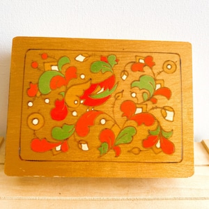 Peut inclure: Boîte rectangulaire en bois avec un motif décoratif floral et d'oiseaux. Le motif présente des accents rouges, verts et blancs sur un fond brun clair. La boîte est posée sur une surface en bois.