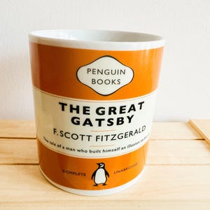 Può includere: Tazza in ceramica con il design della copertina de "Il Grande Gatsby" di F. Scott Fitzgerald. La tazza ha uno schema di colori arancione e bianco con il logo Penguin Books e un'illustrazione di pinguino. Il testo include il titolo e l'autore del libro.