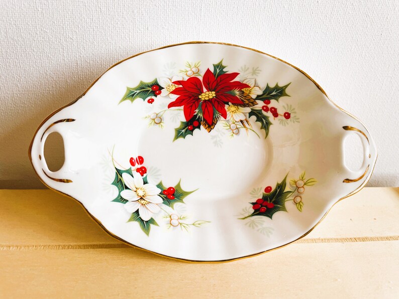 K&ouml;nnte beinhalten: Eine wei&szlig;e Porzellanservierschale mit goldener Verzierung und einem weihnachtlichen Blumendesign. Die Schale zeigt rote Weihnachtssterne, gr&uuml;ne Stechpalmenbl&auml;tter und wei&szlig;e Bl&uuml;ten.