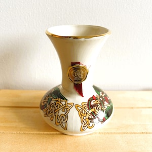 Puede incluir: Un jarrón de cerámica color crema con borde dorado y base bulbosa. El jarrón presenta diseños de nudos celtas en oro, verde y rojo. Un sello dorado con una cinta roja está adherido al jarrón.