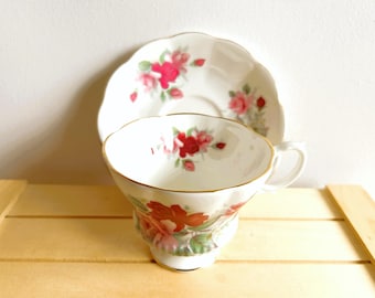Puede incluir: Un juego de taza y platillo blancos con un diseño floral. La taza y el platillo tienen un borde festoneado y ribete dorado. El diseño floral presenta rosas rojas y rosas con hojas verdes. La taza tiene un asa.