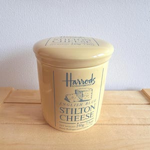 May include: A light yellow ceramic jar with a lid, featuring the text "Harrods Knightsbridge" and "English Blue Stilton Cheese." The jar has a capacity of 7.5 oz and is designed to keep cheese refrigerated.