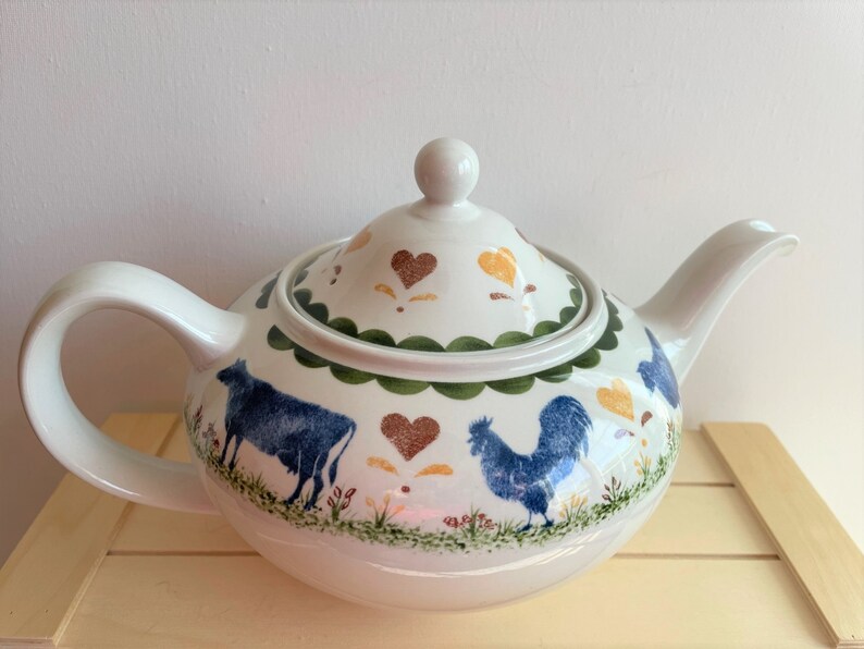 May include: A white teapot with a blue rooster, cow, and green and brown floral designs. The teapot has a lid with a heart design.