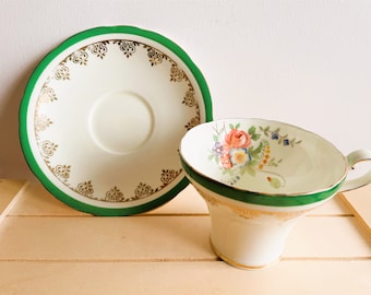 May include: A white teacup and saucer set with a green rim and gold floral pattern. The saucer has a gold and white geometric pattern.