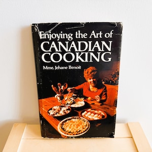Może przedstawiać: Książka kucharska w stylu vintage zatytułowana "Enjoying the Art of Canadian Cooking" autorstwa Mme. Jehane Benoit. Okładka przedstawia kobietę w czerwonej sukience przy stole z różnymi potrawami. Książka leży na drewnianej powierzchni.
