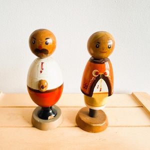 Könnte beinhalten: Zwei bemalte Holzfiguren. Eine hat einen braunen Kopf, ein weißes Hemd und einen roten Rock. Die andere hat einen braunen Kopf, einen orangefarbenen Umhang und einen gelben Rock. Beide stehen auf Holzsockeln.