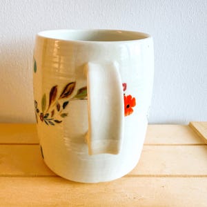 Op de afbeelding: Een witte keramische mok met een gebogen handvat, versierd met bloemmotieven in rood, groen en bruin. De mok staat op een licht houten oppervlak, met een witte muur op de achtergrond.
