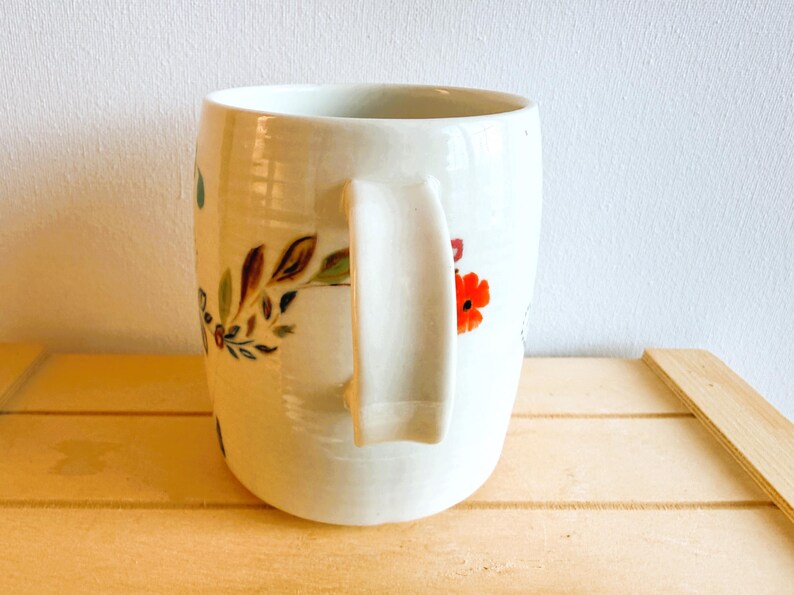 Op de afbeelding: Een witte keramische mok met een gebogen handvat, versierd met bloemmotieven in rood, groen en bruin. De mok staat op een licht houten oppervlak, met een witte muur op de achtergrond.
