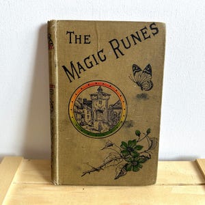 Pode incluir: Um livro vintage intitulado "The Magic Runes" com uma capa marrom. A capa apresenta uma ilustração colorida de um edifício com uma torre de relógio e uma borboleta.