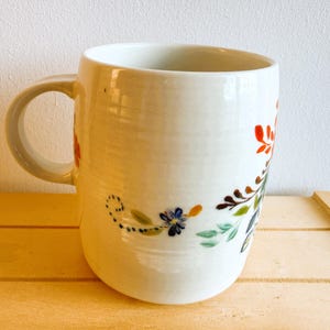 Op de afbeelding: Witte keramische mok met een gebogen handvat, versierd met een bloemenontwerp in rood, blauw, groen en bruin. De bloemen en bladeren zijn met de hand geschilderd. De mok staat op een houten oppervlak.