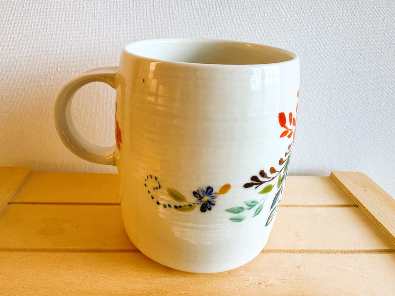 Op de afbeelding: Witte keramische mok met een gebogen handvat, versierd met een bloemenontwerp in rood, blauw, groen en bruin. De bloemen en bladeren zijn met de hand geschilderd. De mok staat op een houten oppervlak.