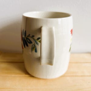 Op de afbeelding: Cr&egrave;mekleurige keramische mok met een gebogen handvat. De mok is versierd met kleurrijke bloemen- en bladerontwerpen in rood, blauw en groen. De mok staat op een lichtbruin houten oppervlak. De mok is ongeveer 10 cm hoog.