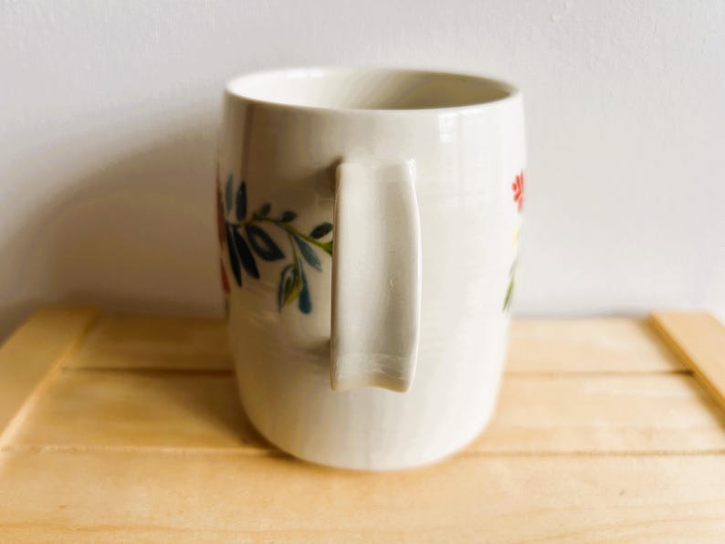 Op de afbeelding: Cr&egrave;mekleurige keramische mok met een gebogen handvat. De mok is versierd met kleurrijke bloemen- en bladerontwerpen in rood, blauw en groen. De mok staat op een lichtbruin houten oppervlak. De mok is ongeveer 10 cm hoog.