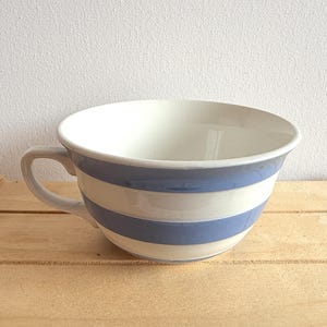 May include: A white ceramic soup bowl with a blue and white striped pattern. The bowl has a handle and is set on a wooden surface. The bowl is empty.