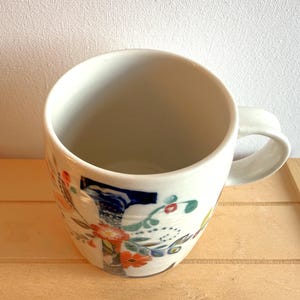 Op de afbeelding: Een witte keramische mok met een bloemenontwerp in oranje, blauw en groen. De mok heeft een afgeronde vorm en een gebogen handvat. De mok staat op een lichtbruin houten oppervlak.