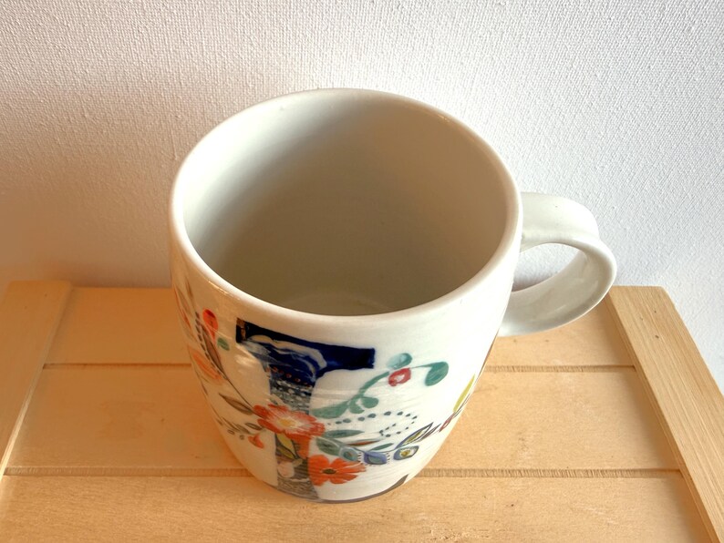 Op de afbeelding: Een witte keramische mok met een bloemenontwerp in oranje, blauw en groen. De mok heeft een afgeronde vorm en een gebogen handvat. De mok staat op een lichtbruin houten oppervlak.