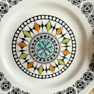 May include: A white ceramic plate with a central design featuring a blue, green, and orange floral pattern. The plate's border has a repeating black and white geometric design. This decorative plate is suitable for serving food.