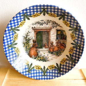 May include: A blue and white gingham patterned ceramic bowl featuring a cottage scene with a door and rabbits. The bowl has a floral border and is ideal for serving soup or salad.