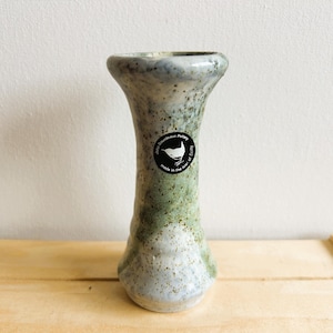 May include: A small, white and green ceramic vase with a speckled glaze. The vase has a round base and a narrow neck. It has a black sticker on the side with the text "John Downham Pottery" and a bird design.