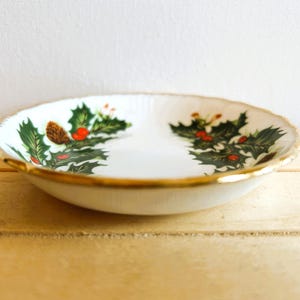 K&ouml;nnte beinhalten: Eine wei&szlig;e Keramikschale mit goldfarbenem Rand und einem Design aus Stechpalme und Beeren. Die Schale befindet sich auf einer Holzoberfl&auml;che.