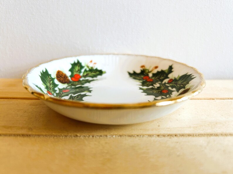 K&ouml;nnte beinhalten: Eine wei&szlig;e Keramikschale mit goldfarbenem Rand und einem Design aus Stechpalme und Beeren. Die Schale befindet sich auf einer Holzoberfl&auml;che.