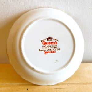 K&ouml;nnte beinhalten: Wei&szlig;e Keramik-Untertasse mit rotem und goldenem Design. Die Untertasse tr&auml;gt den Text "Estd 1875 Queen's Fine Bone China Made in England Rosina China Co. Ltd Yuletide 19" auf der Unterseite.