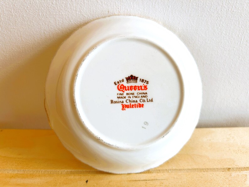 K&ouml;nnte beinhalten: Wei&szlig;e Keramik-Untertasse mit rotem und goldenem Design. Die Untertasse tr&auml;gt den Text "Estd 1875 Queen's Fine Bone China Made in England Rosina China Co. Ltd Yuletide 19" auf der Unterseite.