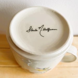 Op de afbeelding: Witte keramische mok met een handvat, met een bloemenontwerp en de handtekening "Starla M. Halfmann" in zwart schrift op de bodem. De mok staat op een houten oppervlak.