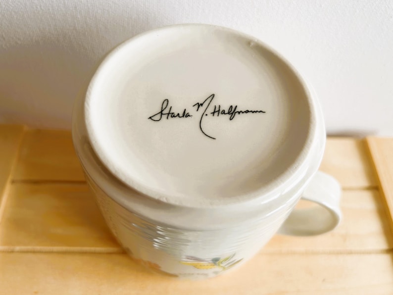 Op de afbeelding: Witte keramische mok met een handvat, met een bloemenontwerp en de handtekening "Starla M. Halfmann" in zwart schrift op de bodem. De mok staat op een houten oppervlak.