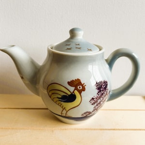 Puede incluir: Una tetera de cerámica azul y blanca con un diseño de gallo. El gallo es amarillo, marrón y negro. La tetera tiene una tapa con una pequeña asa.