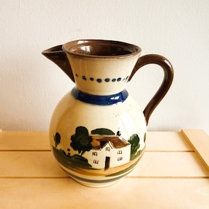 May include: A ceramic jug with a rounded body and a spout. The jug features a hand-painted landscape scene with a house, trees, and a blue band. The handle and rim are brown, and the body is cream-coloured.