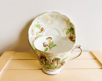 Op de afbeelding: Een wit theekopje en schotel met een bloemenpatroon. Het patroon heeft groene bladeren en roze en witte bloemen. Het kopje en de schotel hebben een gouden rand.