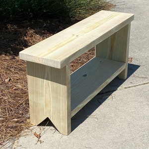 May include: A light-colored wooden bench with a lower shelf. The bench is made of unfinished wood and has a simple, rectangular design. The legs have a decorative cut-out. The bench is approximately 48 inches long.