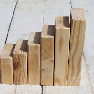 May include: Six unfinished wooden blocks of varying heights arranged in a stepped pattern. The blocks are rectangular, with visible wood grain and knots. The natural wood color contrasts with the white wooden surface they rest on.