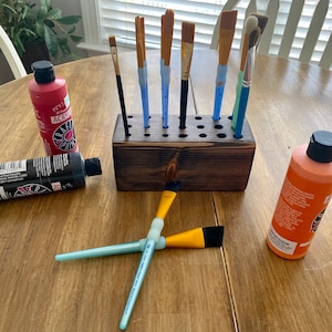 May include: A wooden art brush holder with several paint brushes of various sizes and shapes. Bottles of red, black, and orange acrylic paint are scattered around the holder. The brushes have blue, green, and yellow handles.