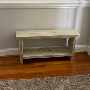 May include: A light-colored wooden bench with a lower shelf. The bench is made of unfinished wood and has a simple, rectangular design. It is placed against a light gray wall and sits on a hardwood floor.