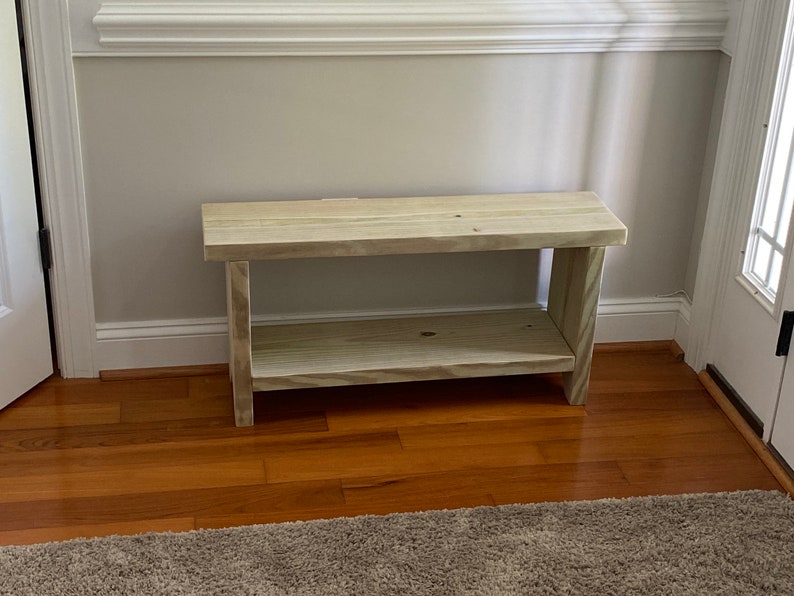 May include: A light-colored wooden bench with a lower shelf. The bench is made of unfinished wood and has a simple, rectangular design. It is placed against a light gray wall and sits on a hardwood floor.