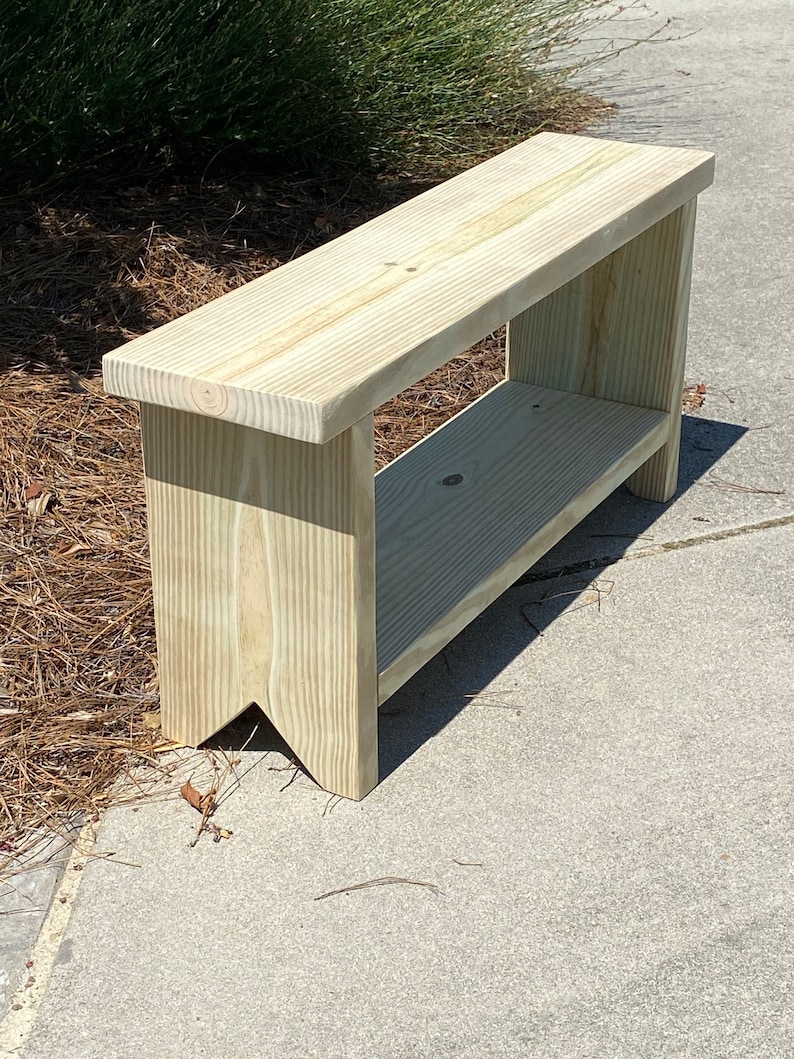 May include: A light-colored wooden bench with a lower shelf. The bench is made of unfinished wood and has a simple, rectangular design. The legs have a decorative cut-out. The bench is approximately 48 inches long.