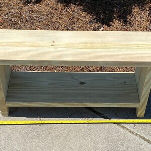 May include: A light-colored wooden bench with a lower shelf. The bench is rectangular and appears to be made of unfinished wood. A yellow measuring tape is placed next to the bench. The bench measures approximately 36 inches long.