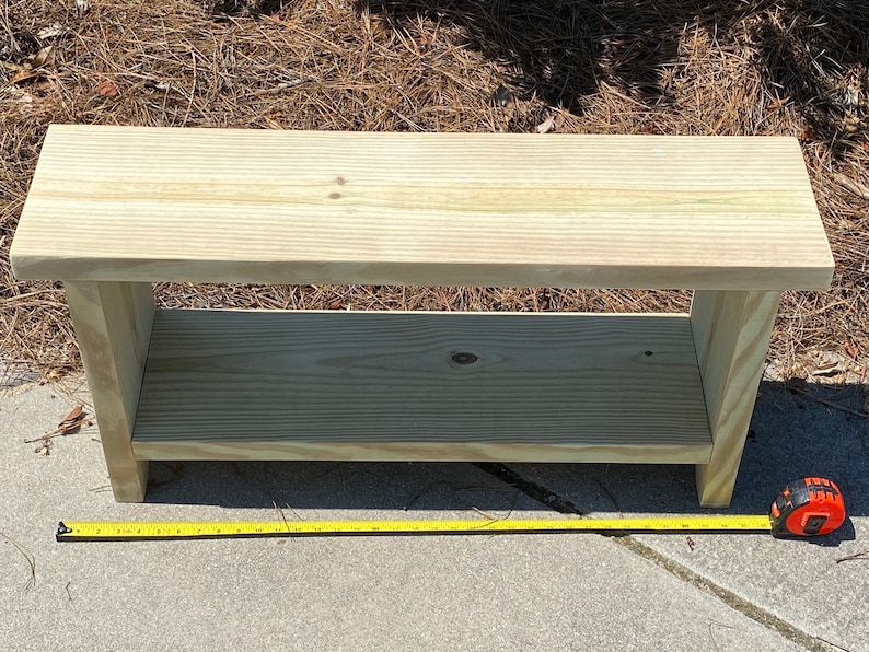 May include: A light-colored wooden bench with a lower shelf. The bench is rectangular and appears to be made of unfinished wood. A yellow measuring tape is placed next to the bench. The bench measures approximately 36 inches long.