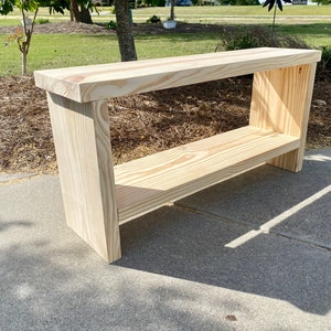 May include: A natural wood console table with a shelf. The table is made of light-colored wood and has a rectangular top. The shelf is positioned below the top, and the legs are thick and sturdy. The table measures approximately 48 inches long, 12 inches wide, and 30 inches high.