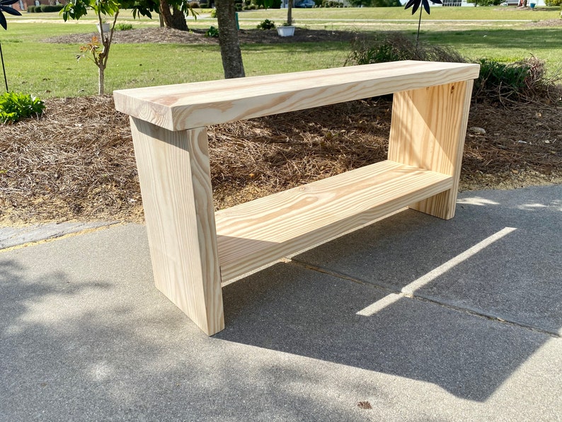 May include: A natural wood console table with a shelf. The table is made of light-colored wood and has a rectangular top. The shelf is positioned below the top, and the legs are thick and sturdy. The table measures approximately 48 inches long, 12 inches wide, and 30 inches high.