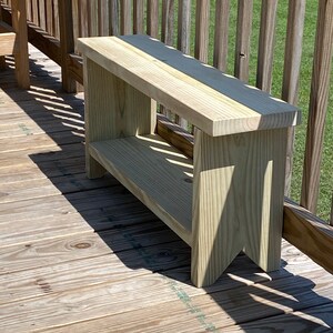 May include: A light-colored wooden bench with a rectangular seat and a lower shelf. The bench has a simple, sturdy design with angled legs. The wood grain is visible, and the bench is placed on a wooden deck.